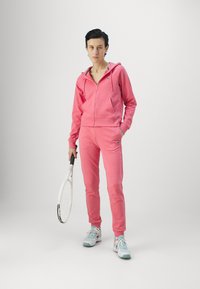 Rausvas treniruočių kostiumas su zip-up gobtuvu ir kelnėmis, pagamintas iš minkštos medžiagos. Modelis laiko teniso raketę, vilkėdamas šviesiai mėlynus sportinius batus.