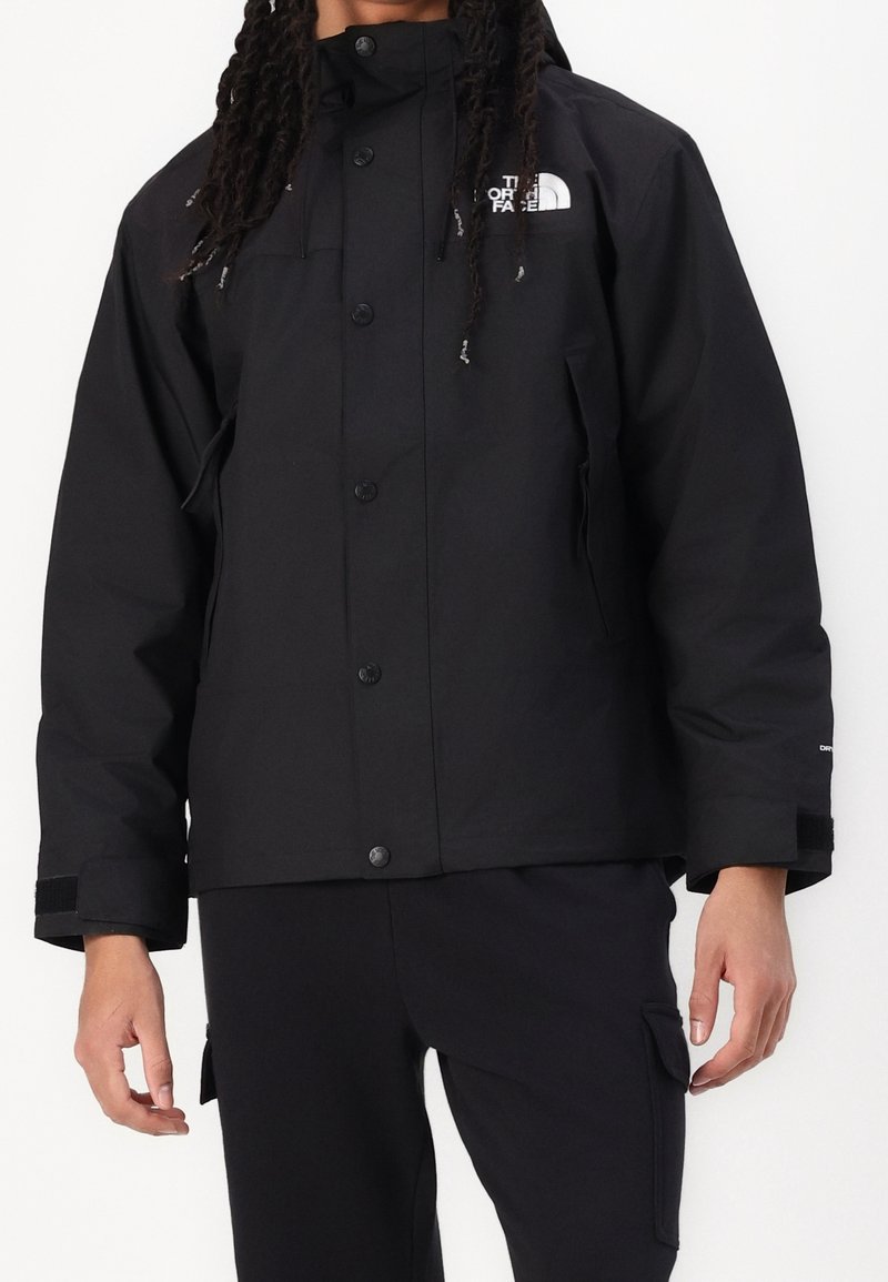 Personne portant une veste noire North Face à fermeture par boutons et un pantalon noir avec poches cargo sur un fond blanc uni.