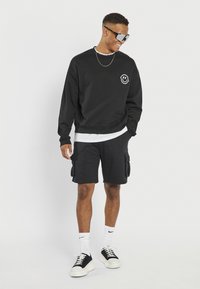 Svart crewneck sweatshirt med frontlogo, kombinert med svarte cargo shorts, hvit undertrøye, hvite sokker og svarte sneakers.