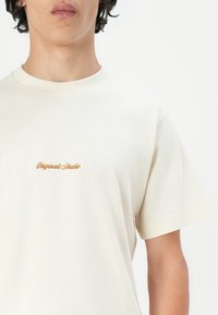 Tricou din bumbac crem cu guler rotund și mâneci scurte, având brodat textul "Originals Studio" în brun deschis pe piept.