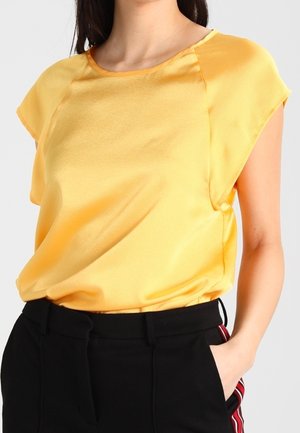 Mujer con blusa de satén amarilla de manga corta metida dentro de pantalones negros con rayas laterales rojas y blancas, con la mano en el bolsillo.