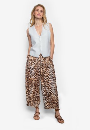 Vrouw staat met handen in zakken, draagt een mouwloos wit topje met knopen, wijde broek met luipaardprint en bruine sandalen met bandjes.