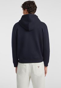 Marineblaues Kapuzen-Sweatshirt mit lockerem Schnitt, ausgestattet mit gerippten Bündchen und Saum. Kombiniert mit weißen Jeans, akzentuiert durch ein Logo-Patch auf der Rückseite.