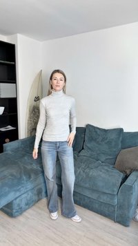 Szürke garbó pulóver világoskék, bővülő farmernadrággal. Hátterében kényelmes tealdzseki kordbársony kanapé, fehér fal és világos fapadló.