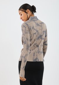 Ribbet langermet topp i beige med blå tie-dye mønster, med en tettsittende design og høy krage, iført med et svart skjørt.