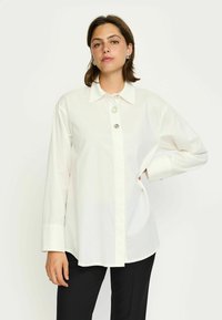 Cămașă albă cu nasturi, din bumbac, cu mâneci lungi, guler ascuțit și un nasture decorativ la gât. Asortată cu pantaloni negri.