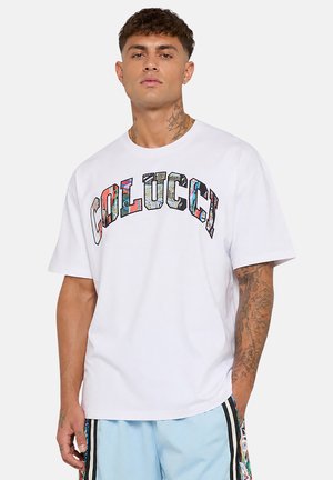 Mann mit Hals- und Armtattoos, der ein weißes Colucci-T-Shirt mit farbigem Logo und hellblauen Shorts mit schwarzen Streifen trägt.