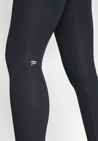 Leggings atletici neri realizzati in tessuto elasticizzato, con una texture liscia e un piccolo logo bianco vicino al ginocchio. Dettagli delle cuciture visibili.
