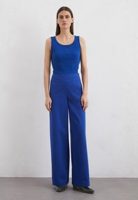 Débardeur bleu côtelé associé à un pantalon large taille haute bleu. Le modèle porte des bottines noires à talons. Tenue simple et unie.