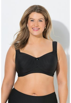 Donna sorridente con capelli fino alle spalle che indossa un reggiseno nero a righe sottili e slip abbinati su uno sfondo chiaro uniforme.