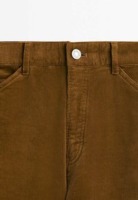 Pantalon en velours côtelé marron avec texture à côtes verticales, fermeture par bouton à la taille et poches latérales. Coutures renforcées le long des coutures.