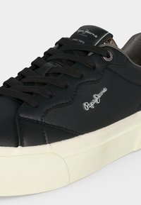 Μαύρο sneaker Pepe Jeans με μαύρα κορδόνια, λευκή σόλα και λογότυπο στη πλευρά και στη γλώσσα, που δείχνει λεπτομερή ραφή και οπές για κορδόνια.