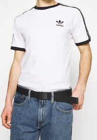 Bílé tričko s krátkým rukávem a černým límečkem a okraji rukávů, na prsou s malým černým logem Adidas, kombinované s modrými džínami.