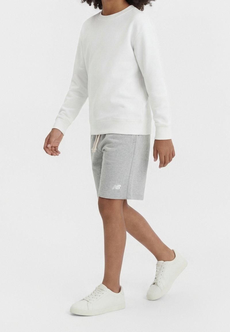 Personne portant une chemise blanche à manches longues, un short gris avec cordon et logo, et des baskets blanches, marchant sur un fond blanc uni.