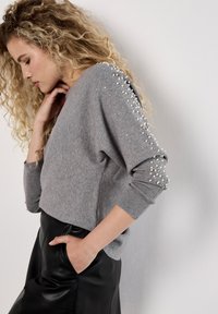 Grau gestrickter Pullover mit lockerer Passform, verziert mit silbernen Perlen entlang der Ärmel, aus strukturiertem Strickstoff und mit gerippten Bündchen. Kombiniert mit einem schwarzen Lederrock.