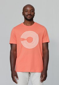 T-shirt en coton corail avec un design graphique à rayures circulaires blanches. Manches courtes et col ras du cou, porté avec un pantalon de couleur claire.