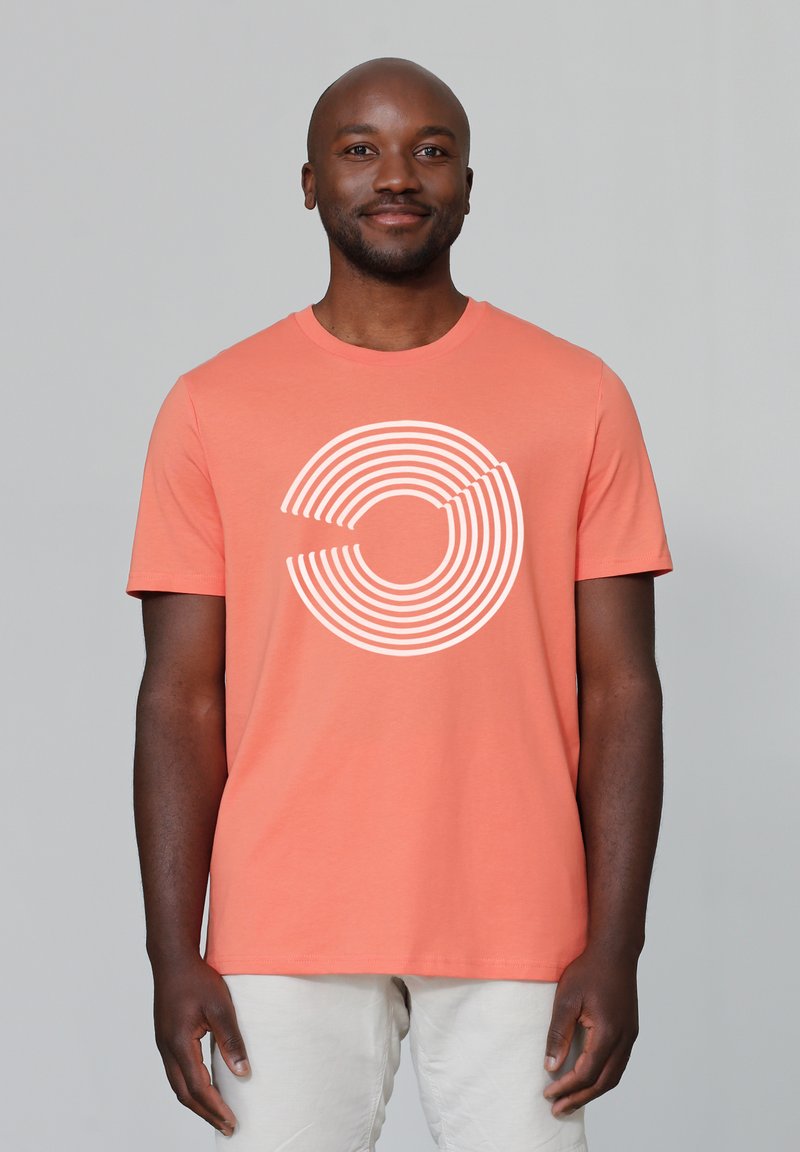 T-shirt en coton corail avec un design graphique à rayures circulaires blanches. Manches courtes et col ras du cou, porté avec un pantalon de couleur claire.