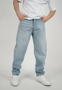 Person, der eine hellblaue, lockere Jeans, weiße Sneakers und ein weißes T-Shirt trägt, mit den Händen in den Taschen, vor einem weißen Hintergrund stehend.