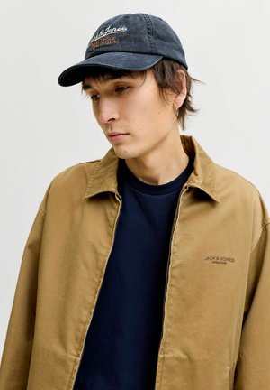 Veste en coton marron avec une fermeture éclair, t-shirt bleu marine en dessous, et casquette bleu marine avec texte brodé en blanc et orange. Le modèle a les cheveux courts.