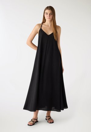 Giovane donna che indossa un abito maxi nero senza maniche con spalline sottili e sandali neri bassi, in piedi contro uno sfondo bianco uniforme.