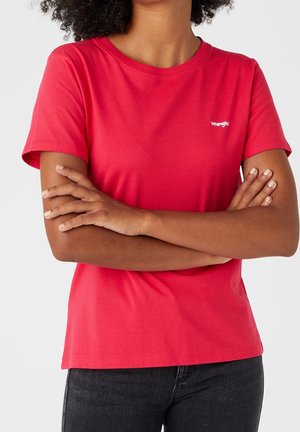 Frau mit verschränkten Armen, die ein rotes Wrangler-T-Shirt und schwarze Jeans trägt, von Hals bis Hüfte vor weißem Hintergrund gezeigt.
