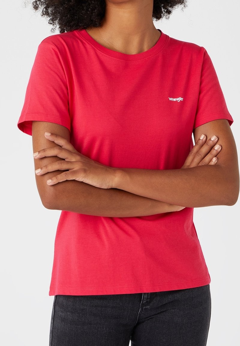 Femme les bras croisés portant un t-shirt rouge Wrangler et un jean noir, montrée du cou aux hanches sur fond blanc.