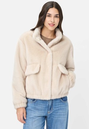 Junge Frau trägt eine hellbeige Kunstpelzjacke mit Fronttaschen und blaue Jeans, steht vor einem schlichten weißen Hintergrund.