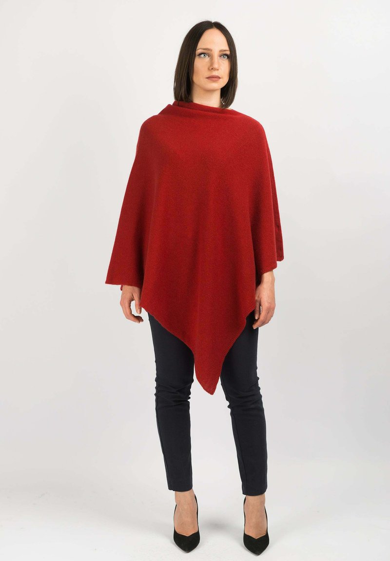 Puncho punainen villa kolmion muotoinen, pehmeä tekstuuri ja rullattu kaulus. Päällä tummat housut ja mustat korkokengät.