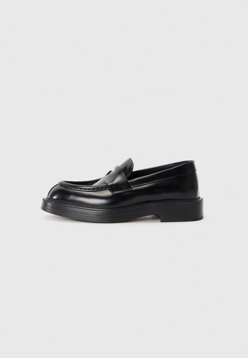 Mocasines de cuero negro con punta redondeada, un diseño cosido en la parte superior y una suela de goma gruesa. Textura suave con acentos mínimos.