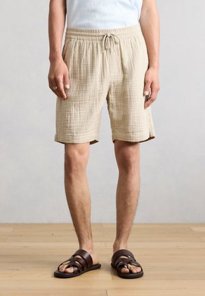 Mężczyzna w beżowych szortach ze sznurkiem i ciemnobrązowych sandałach stoi na drewnianej podłodze na tle jednolitej ściany.