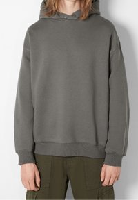 Persona che indossa un ampio hoodie grigio con bottoni a scatto vicino al colletto e pantaloni cargo verde oliva, in piedi contro uno sfondo chiaro.