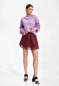 Lavendel gebreid sweater met uitsparingen in de mouwen, gecombineerd met een rok met rode bloemenprint. Model draagt zwarte sandalen met blokhak.