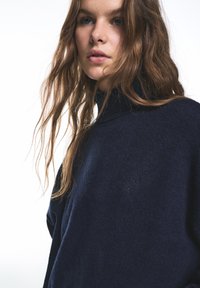 Granatowy sweter z golfem wykonany z miękkiego dzianinowego materiału, o luźnym kroju i subtelnym akcentem logo na piersi.