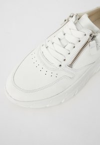 Baskets blanches avec dessus en cuir texturé, perforations prononcées, lacets plats et une fermeture éclair latérale. Semelle épaisse blanche avec un motif de bande de roulement distinctif.