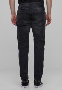 Jeans de mezclilla negra con corte slim, diseño de cinco bolsillos y cierre con cremallera. Presenta un parche de cuero en la parte trasera de la cintura y un detalle de cadena en el costado.