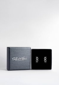 Orecchini a forma di cuore in argento esposti in una scatola foderata in velluto nero con un esterno in argento texturizzato, con il nome del marchio in bianco.