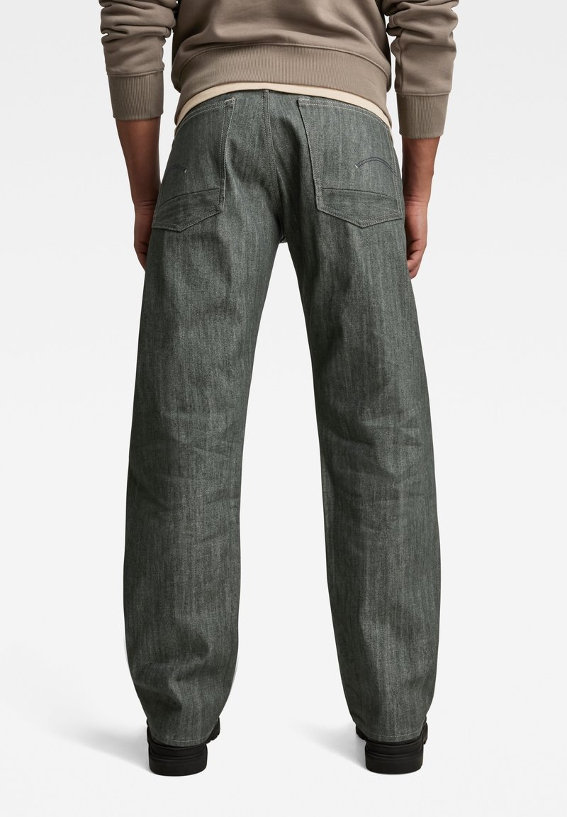 Persoon in losse, donkergrijze jeans en een beige sweatshirt, gezien van achteren tegen een witte achtergrond.