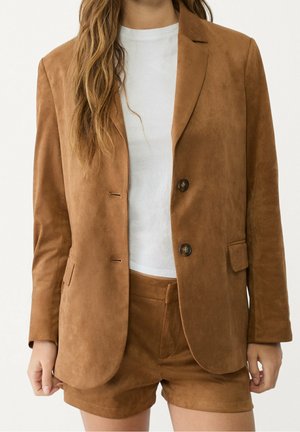 Femme portant un blazer en daim marron à deux boutons et un short assorti par-dessus un t-shirt blanc uni, debout devant un fond blanc.