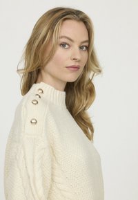 Pull en maille couleur crème avec un col montant, arborant un motif en torsades et de gros boutons décoratifs sur une épaule.