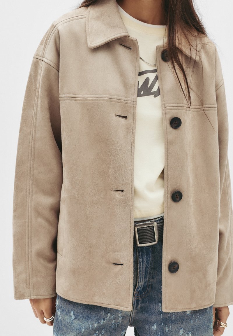 Femme portant une veste beige boutonnée sur un t-shirt blanc à motif graphique, associée à un jean bleu déchiré et une ceinture noire avec une boucle carrée.