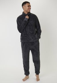 Un jeune homme se tient pieds nus, portant un pull en peluche gris foncé et un pantalon assorti, souriant et tenant le col de son pull.