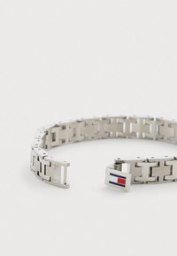 Zilveren metalen armband met in elkaar grijpende rechthoekige schakels, voorzien van een sluiting die wordt geaccentueerd door een logo met rode, witte en blauwe details.