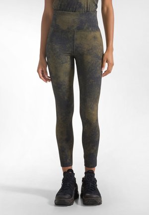 Kvinde iført højtaljede, grønne og sorte tie-dye leggings og sorte træningssko, der står mod en ensfarvet lys baggrund.