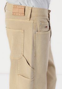 Beige katoenen broek met een gestructureerde afwerking, met meerdere zakken en een leren label met de tekst "TOMMY JEANS." Subtiele logo-accenten zichtbaar.