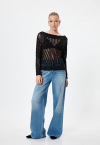 Zwarte gebreide trui met een los, open structuur en brede bateauhals, gecombineerd met lichtblauwe wijde jeans en zwarte schoenen.