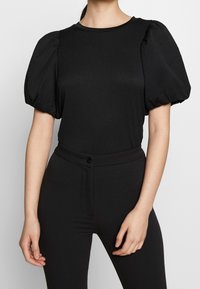 Top nero con maniche a puff realizzato in tessuto strutturato con scollatura rotonda, abbinato a pantaloni neri a vita alta con dettaglio a bottone.