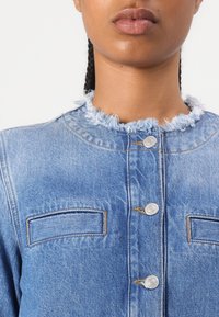 Veste en denim bleu avec un col effiloché, des boutons en métal argenté et deux poches poitrine. Présente des coutures contrastées et une coupe décontractée.