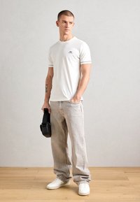 T-shirt di cotone bianca con bordi verdi, jeans grigio chiaro e sneakers bianche; tiene in mano una borsa rotonda nera, in piedi su un pavimento di legno.