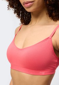 Γυναίκα με σγουρά μαλλιά που φοράει ένα απαλό κοραλί ροζ σουτιέν με λεπτές τιράντες και χωρίς ορατά ραφές ή ενίσχυση, σε λευκό φόντο.