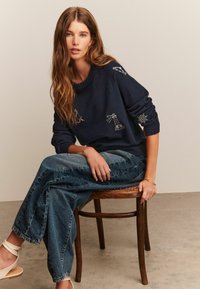 Mörkblå tröja med broderade nautiska motiv, tillsammans med ljusblå vida denimjeans. Sitter på en trästol, minimalistisk miljö.